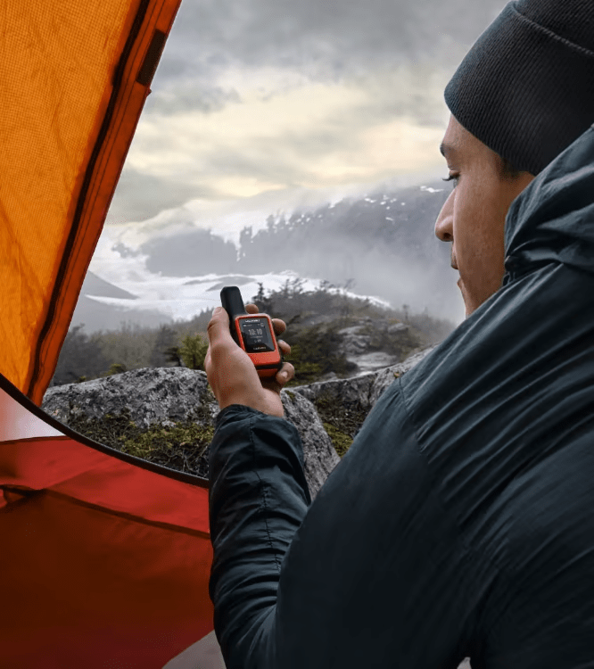 Camper in a winter jacket in a tent doorway holding a GPS communicator, snowy mountains behind