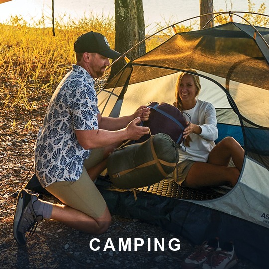 Couple unpacking a sleeping bag at a backpacking tent by a lake at sunset, caption CAMPING