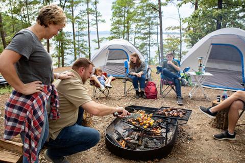 Family cooking over a fire ring at a lakeside campsite with camp chairs and tents