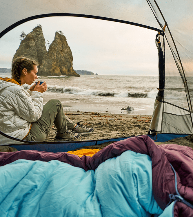 Camper sitting in an open tent with sleeping bags, looking out over a rocky coastline