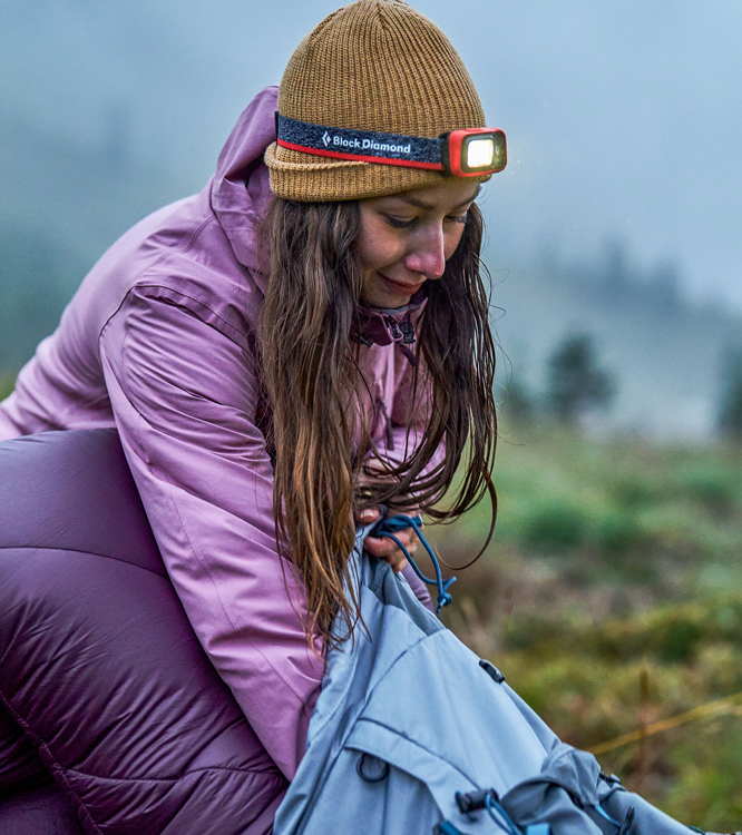 Camper wearing a headlamp over a beanie, packing a bag at dusk in the fog