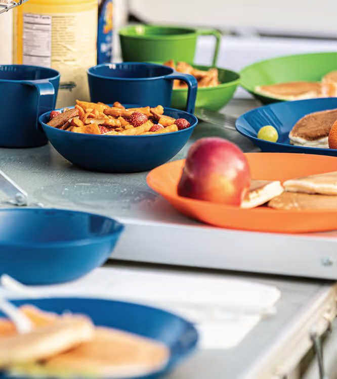 Colourful camp plates, bowls and mugs with breakfast laid out on a camp kitchen table